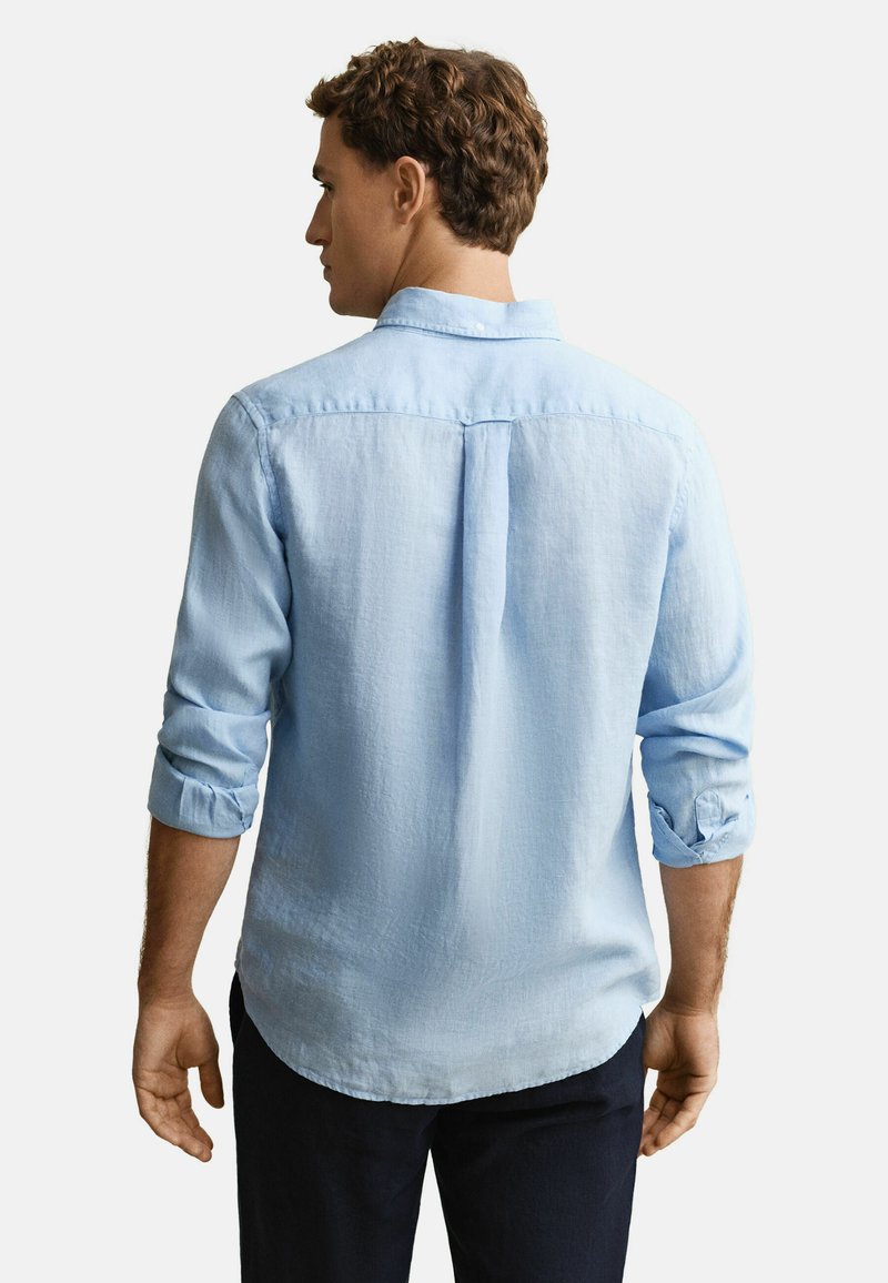 Mann von hinten gesehen, trägt ein hellblaues Hemd mit langen Ärmeln, die hochgekrempelt sind, und eine dunkle Hose vor weißem Hintergrund.