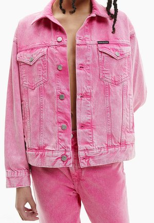 Person with braided hair wears unbuttoned pink denim jacket and matching jeans, showing bare chest, brand tag visible on jacket pocket.