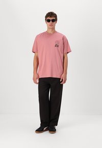Lichtroze katoenen t-shirt met een grafische print op de linker borst, gecombineerd met losse zwarte broeken en zwarte schoenen. Model draagt een zonnebril.
