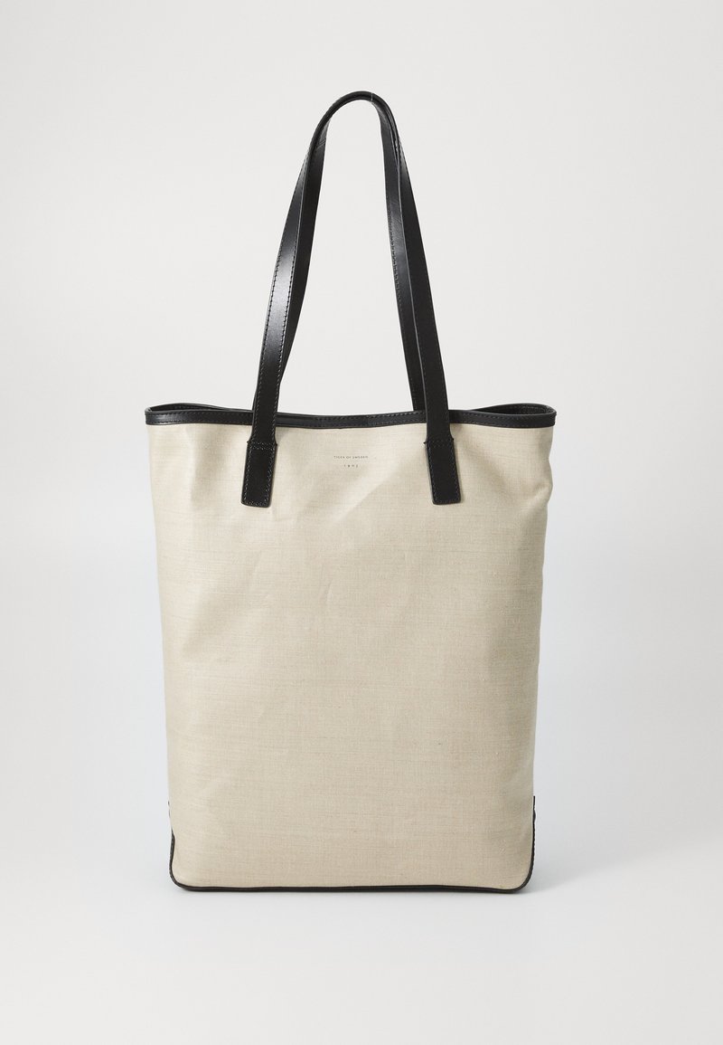 Canvas-Tragetasche in Beige mit schwarzen Lederausschnitten und -griffen. Rechteckige Form, strukturierte Oberfläche, minimalistisches Design, ohne sichtbaren Verschluss.