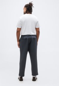 Άνδρας με κοτσίδες δεμένες πίσω στέκεται με την πλάτη προς τα εμπρός, φοράει λευκό μπλουζάκι, σκούρο παντελόνι με πίσω τσέπες, λευκές κάλτσες και καφέ παπούτσια.