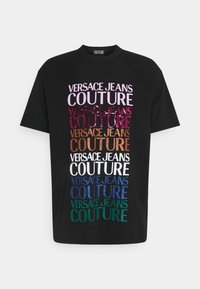 Sort bomulds-T-shirt med stablet tekst "VERSACE JEANS COUTURE" i forskellige farver og glitrede finish, der viser et moderne, dristigt design.