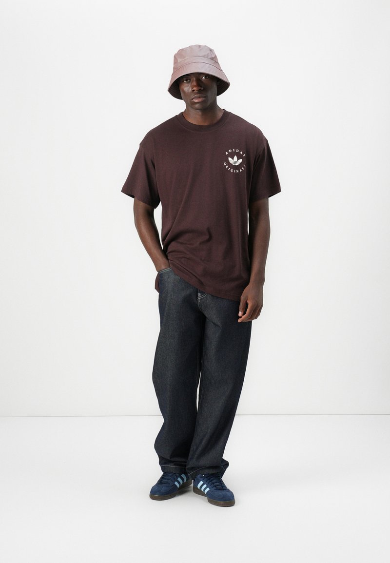 T-shirt in cotone marrone con grafica frontale, jeans in denim scuro, cappello a secchiello rosa chiaro e sneakers blu Adidas con strisce azzurre.