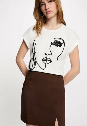 Mladá žena nosící bílé tričko s černým abstraktním obličejem z linek a tmavě hnědou sukni s vysokým pasem s knoflíkovými detaily.