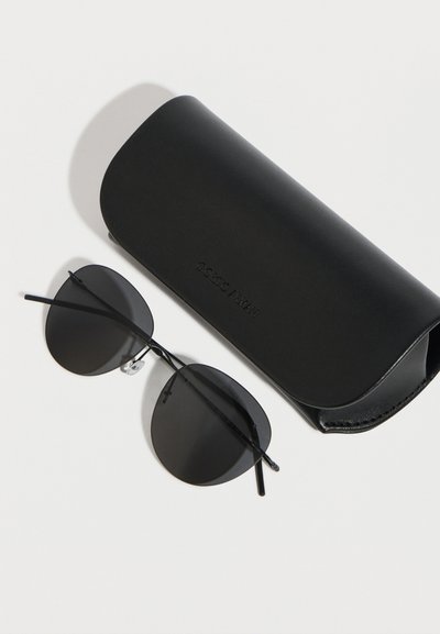 Μαύρα στρογγυλά γυαλιά ηλίου με λεπτά πλαίσια δίπλα σε μια μαύρη θήκη προστασίας με το λογότυπο Giorgio Armani, πάνω σε λευκή επιφάνεια.