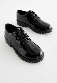 Chaussures en cuir verni noir avec un bout arrondi, un laçage, et une semelle en caoutchouc texturée présentant des crampons proéminents pour une meilleure adhérence.