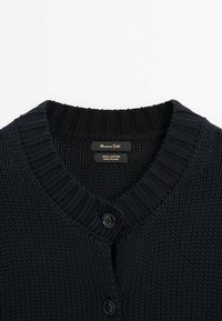 Marineblauwe katoenen gebreide cardigan met een geribde kraag, twee zwarte knopen en een label met de tekst "100% katoen, gemaakt in China."