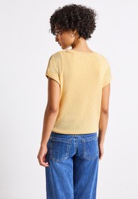 Gul kortärmad texturerad tröja med lös passform, med rund halsringning och uppvikta fållar, tillsammans med blå vida denimjeans.