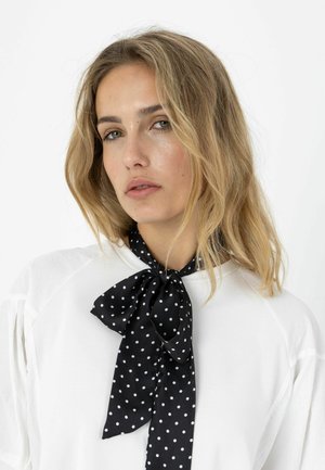 Nő szőke hajjal, fehér felsőt és a nyakában masnira kötött fekete pöttyös sálat visel.