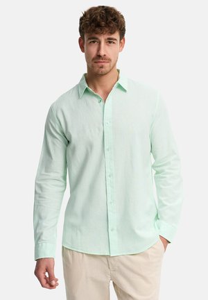 Joven con cabello rizado castaño, vestido con una camisa de manga larga de color verde claro abotonada y pantalones beige, de pie frente a un fondo blanco.