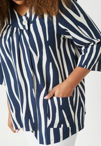 Top a strisce navy e crema con chiusura a bottoni, maniche corte e tasca laterale, caratterizzato da una vestibilità ampia e un tessuto strutturato.