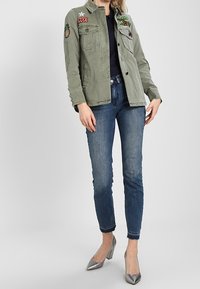 Oliivinvihreä takki, jossa on brodeerattuja kuvioita, yhdistettynä tummansinisiin kapeisiin farkkuihin ja hopeisiin korkokenkiin. Rakenteeltaan teksturoitu kangas ja mittatilaustyönä tehty istuvuus.