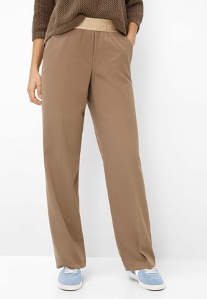 Persona con pantaloni marroni larghi a vita elastica e scarpe da ginnastica blu, con la mano in tasca, in piedi contro uno sfondo bianco.