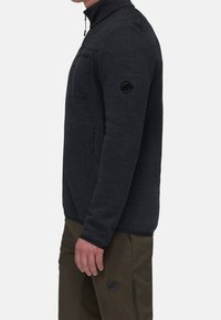 Giacca in pile di colore grigio scuro con colletto alto, tasca frontale con zip e patch con logo sulla manica. Tessuto testurizzato con maniche aderenti.