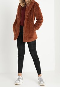 Rostfärgad fuskpälsjacka med bred krage och enkel knappknäppning, i kombination med svarta slim fit-jeans och vita sneakers.