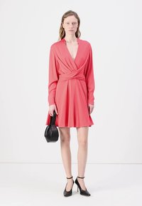 Coral wrap dress with long sleeves, a deep V-neck, and flared skirt; paired with a small black bag and black high-heeled shoes.