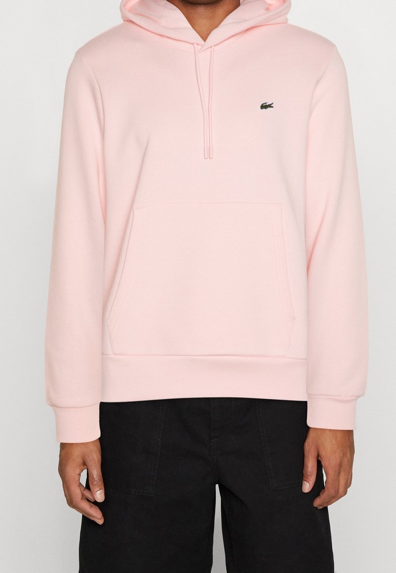 Personne portant un sweat à capuche rose clair avec poche à l'avant et petit logo, associé à un pantalon noir, montré du torse aux hanches.