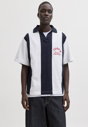 Personne portant un maillot de rugby à rayures verticales bleu marine et blanc avec un texte rouge sur le côté gauche de la poitrine et un jean foncé, debout devant un fond uni.