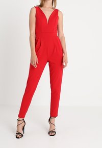 Jumpsuit rosso senza maniche con profondo scollo a V, vita aderente e gambe affusolate, realizzato in tessuto morbido. Presenta pieghe e tacchi neri con cinghie.