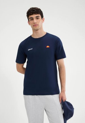 Hombre joven con camiseta azul marino de ellesse y pantalones gris claro, sosteniendo una gorra azul marino a juego en su mano derecha.