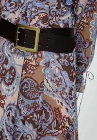 Široké šaty s potiskem, které prezentují vířící paisley vzory v modré, hnědé a růžové barvě, stáhnuté v pase černým koženým páskem se zlatou sponou.