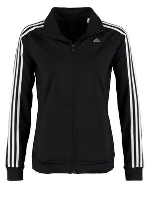 Persoon draagt een zwart Adidas trainingspak met witte strepen en witte sneakers, staand tegen een effen achtergrond.