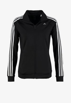 Personne portant un survêtement Adidas noir avec des bandes blanches et des baskets blanches, debout contre un fond uni.