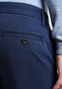 Navy blue trousers with a smooth texture, featuring a back pocket and a button detail. Fabric appears durable and offers structured lines.