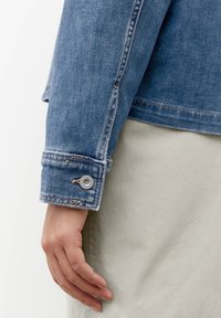 Giubbotto di jeans con lavaggio blu, dotato di polsini con bottoni, cuciture a vista e orlo grezzo. Mano appoggiata su un tessuto chiaro sottostante.