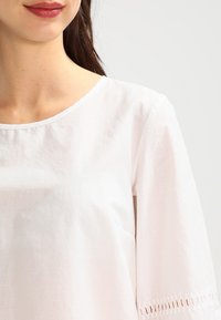 Vit bomullskjorta med rund halsringning, korta ärmar och delikat hålsöm broderi vid ärmsluten. Slät textur och löst sittande.
