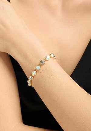 Guldarmbånd med skiftevis klare og iriserende runde ædelsten båret på en underarm mod en sort ærmeløs beklædningsbaggrund.