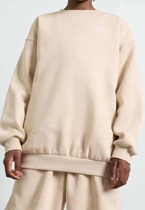 Person iført en beige Nike sweatshirt og matchende bukser, som viser ribbede manchetter og logoet på øverste venstre bryst.