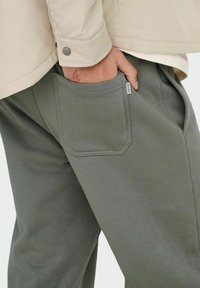 Les pantalons de survêtement gris présentent une finition texturée, des poches latérales et une poche à rabat distinctive à l'arrière. Portés avec une veste beige.