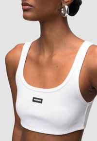 Top corto bianco a costine con spalline larghe e scollatura arrotondata, caratterizzato da un'etichetta nera con testo bianco centrato sulla parte anteriore.