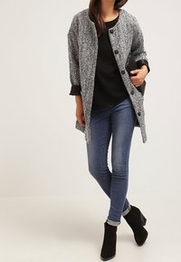 Cappotto grigio e nero con texture e chiusura a bottone, indossato sopra un top nero e jeans blu, abbinato a stivaletti neri con tacco.
