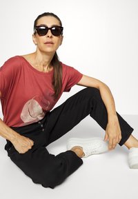 Camisa curta vermelha com um gráfico abstrato, calças pretas de perna reta e ténis brancos. Os acessórios incluem óculos de sol escuros.