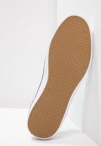 Baskets blanches avec une semelle en gomme texturée, présentant un motif géométrique. Le côté révèle un design lisse avec un accent rouge contrastant.