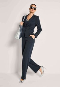 Marineblauer zweiteiliger Anzug mit maßgeschneidertem Sakko und gerade geschnittenen Hosen, kombiniert mit metallisch silbernen High Heels und einer glänzenden silbernen Handtasche.