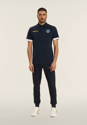 Uomo che indossa una polo blu scuro con logo e pantaloni da jogging abbinati, in piedi contro uno sfondo chiaro e uniforme.