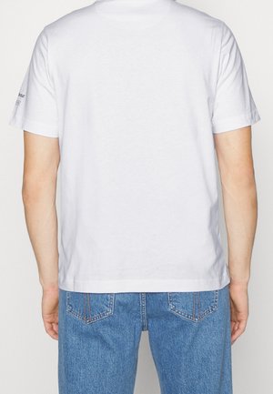 Osoba ubrana w gładki biały t-shirt z krótkim rękawem oraz niebieskie dżinsy, widziana od tyłu na jednolitym tle.