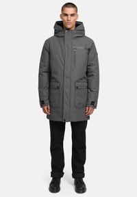 Grå parkajacka med huva, som har stora frampockets, en dragkedjeficka på bröstet och textilstruktur. Bärs över svarta byxor och stövlar.