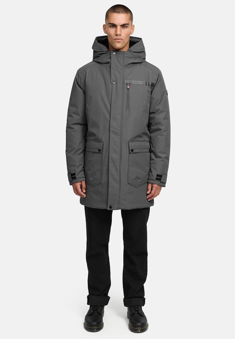 Grå parkajacka med huva, som har stora frampockets, en dragkedjeficka på bröstet och textilstruktur. Bärs över svarta byxor och stövlar.