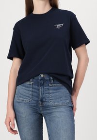 Námořnické bavlněné tričko s krátkým rukávem, s bílým logem "tommy Jeans" na levé části hrudi. Spárováno s džíny s vysokým pasem v modré barvě.