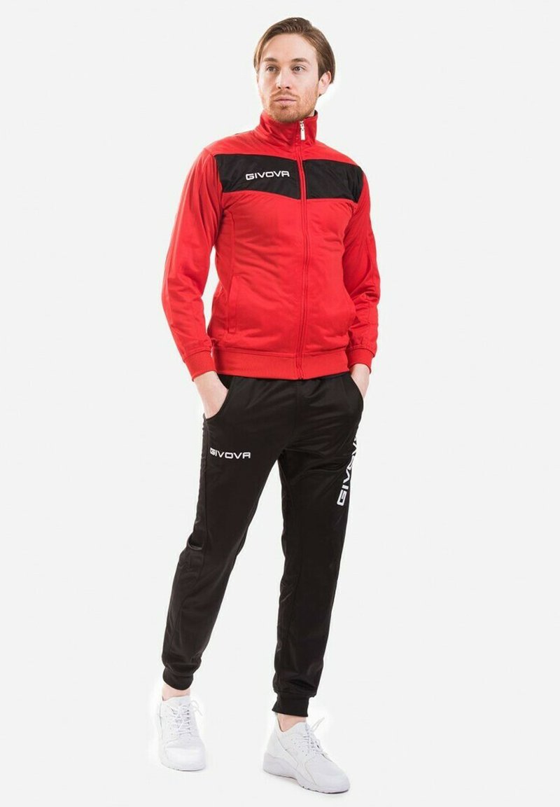 Giacca da track rossa con dettagli neri, colletto alto e chiusura con zip, abbinata a pantaloni neri con logo. Scarpe da ginnastica bianche.