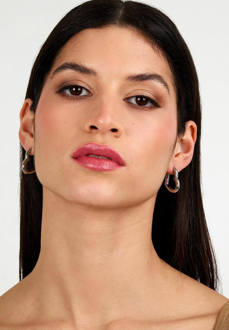 Giovane donna con capelli scuri e lisci indossa orecchini a cerchio d'argento e un trucco sottile con rossetto rosa, guardando direttamente verso la fotocamera.