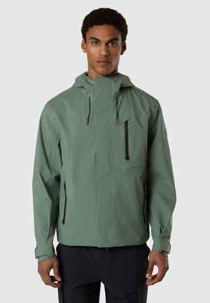 Hombre vestido con chaqueta impermeable con capucha color verde salvia, con bolsillos negros con cremallera y pantalones negros, de pie contra un fondo claro y liso.