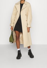 Manteau beige à double boutonnage, avec de larges revers et des manches retroussées, porté sur une robe noire. Bottes noires à plateforme et sac vert.
