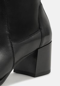 Botină din piele neagră până la gleznă, cu toc pătrat. Detalii de cusătură distincte și o textură netedă. Înălțimea tocului oferă stabilitate și sprijin.