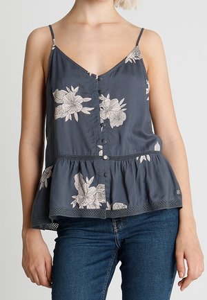 Woman wearing dark blue floral sleeveless buttoned blouse with ruffled hem and blue jeans, standing against plain background.