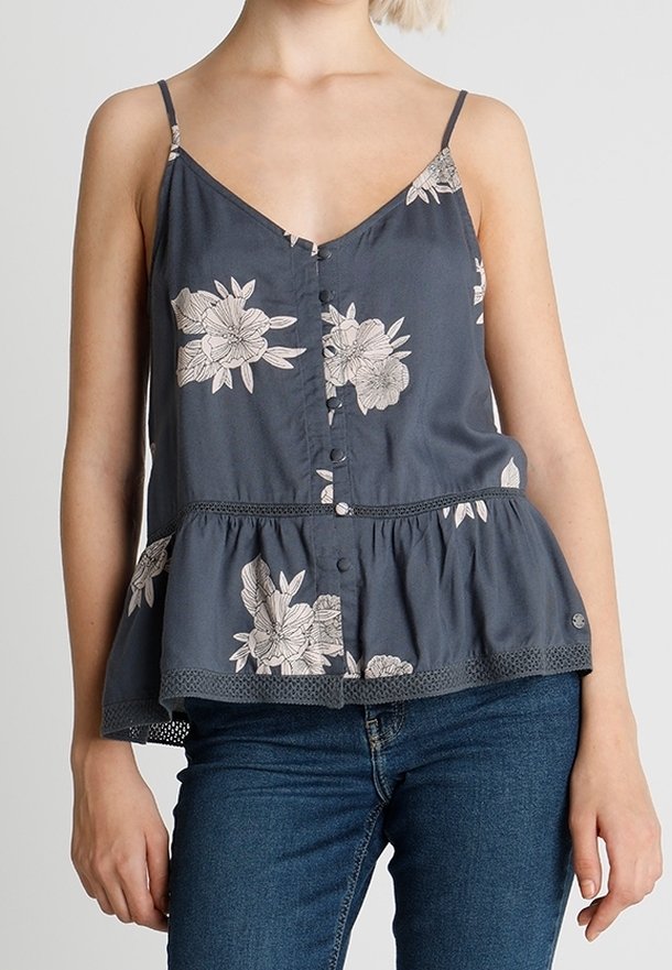 Woman wearing dark blue floral sleeveless buttoned blouse with ruffled hem and blue jeans, standing against plain background.